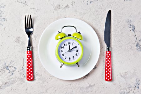 An alarm clock on a white plate between a fork and a knife to symbolize intermittent fasting. An alarm clock on a plate in a table setting