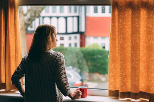 A woman standing indoors looking out a window. 